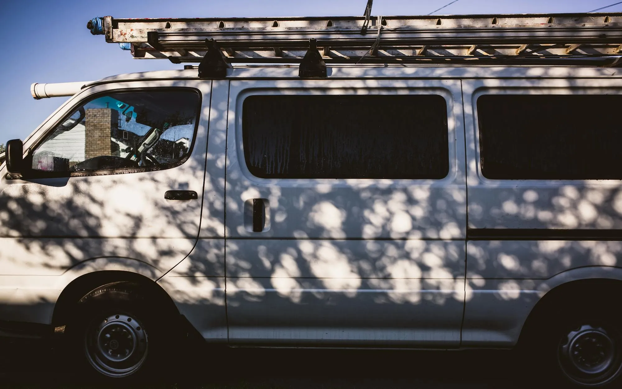 A clean white service van parked under trees, ladder on the roof rack
